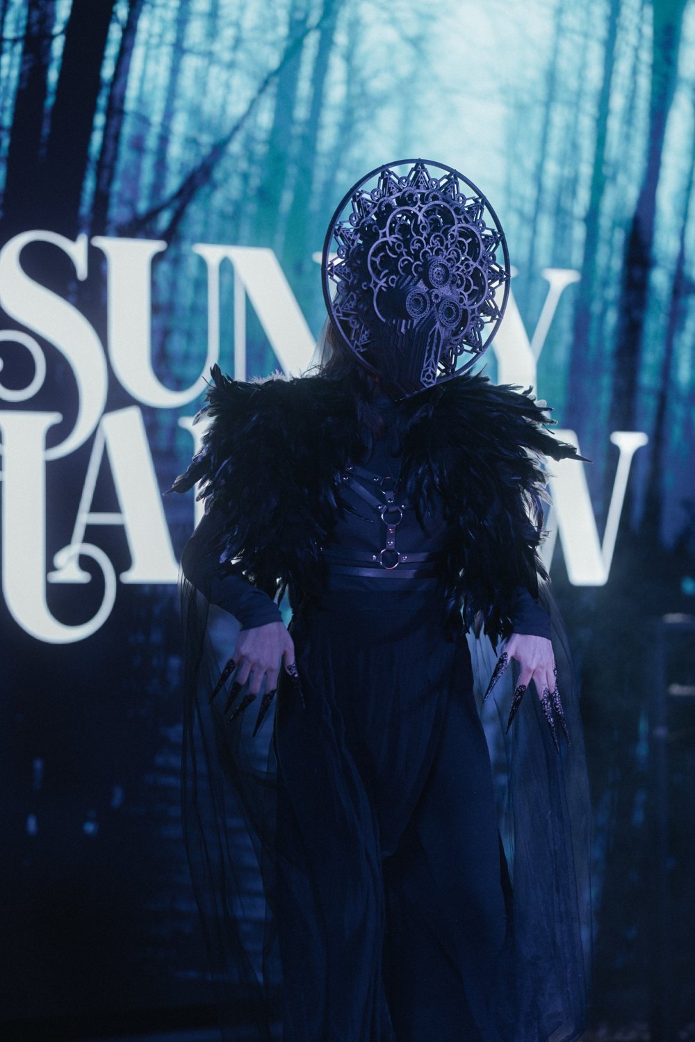 The Temptress: a figure in an ornate black filigree mask and feathered shoulders, with long black claws, standing in a misty blue forest