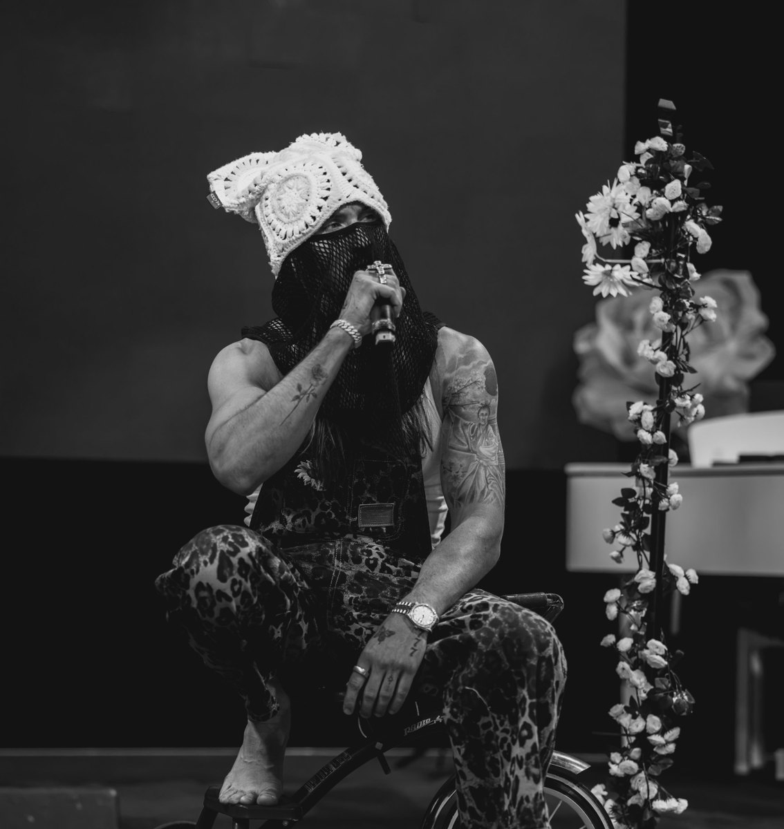 Black and white photo of Sunny on stage in a white crochet hat and black mesh veil, singing into a microphone while perched on a vintage tricycle beside a column of white roses